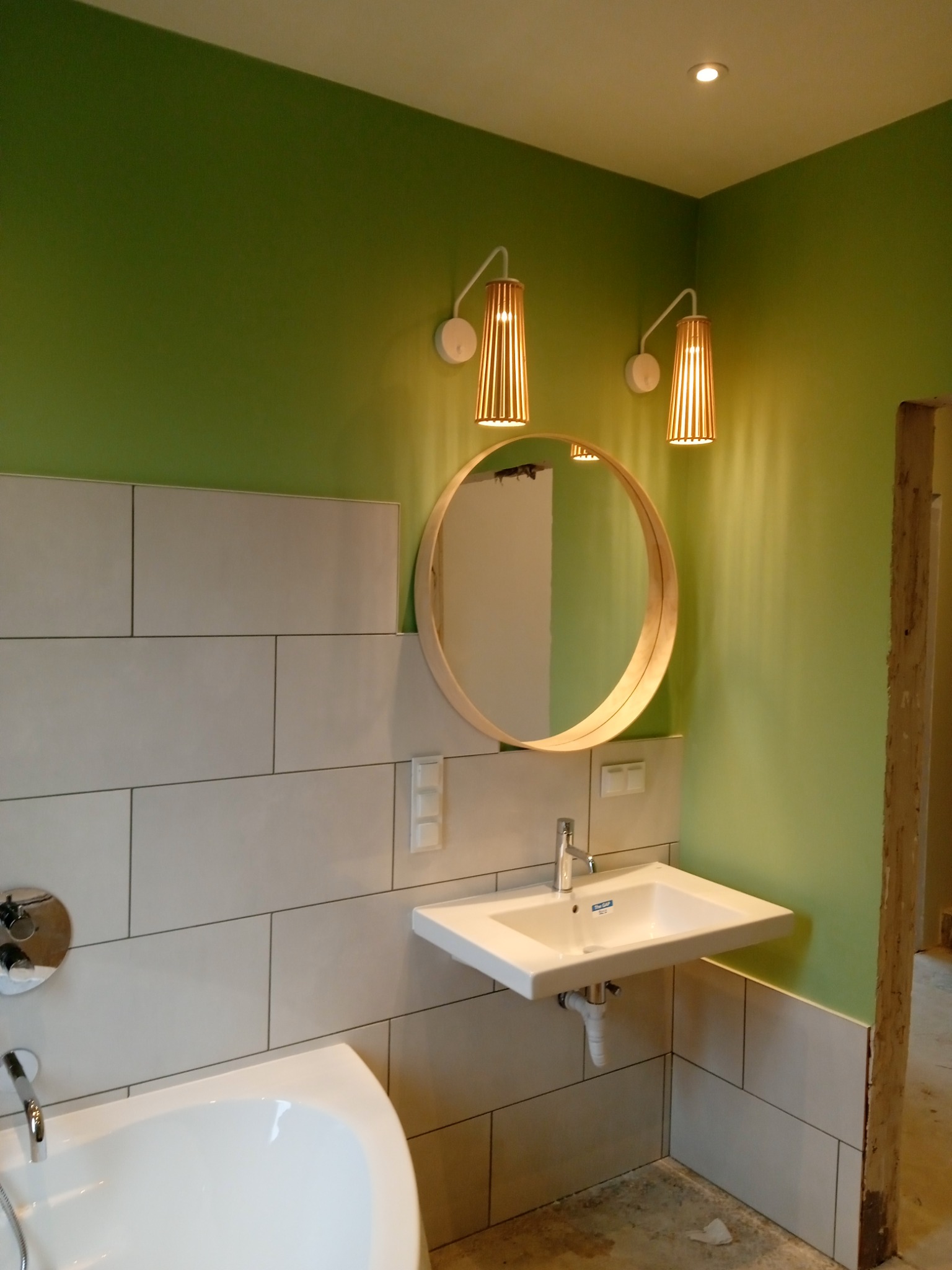 Wykończona łazienka z zielonymi ścianami, białymi płytkami ułożonymi poziomo, okrągłym lustrem z drewnianą ramą, białą umywalką wiszącą i drewnianymi lampami ściennymi z ciepłym światłem.