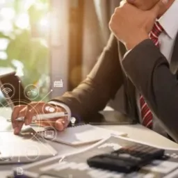 Mężczyzna w garniturze, zamyślony, pracuje na laptopie przy biurku z notatnikiem, kalkulatorem i kubkiem, otoczony ikonami symbolizującymi usługi cyfrowe.