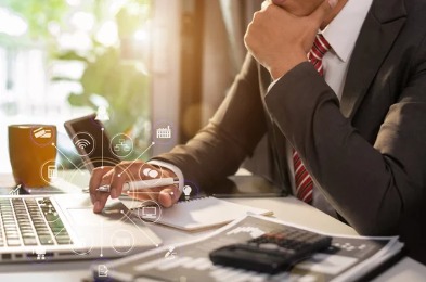 Mężczyzna w garniturze, zamyślony, pracuje na laptopie przy biurku z notatnikiem, kalkulatorem i kubkiem, otoczony ikonami symbolizującymi usługi cyfrowe.