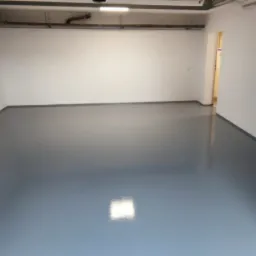 Szara, błyszcząca posadzka epoksydowa w pustym pomieszczeniu z białymi ścianami i widocznymi rurami pod sufitem.