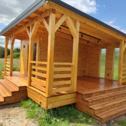 Drewniany domek letniskowy z tarasem i schodami, wykończony na wysoki połysk, z widocznymi lampami sufitowymi i zielonym otoczeniem.