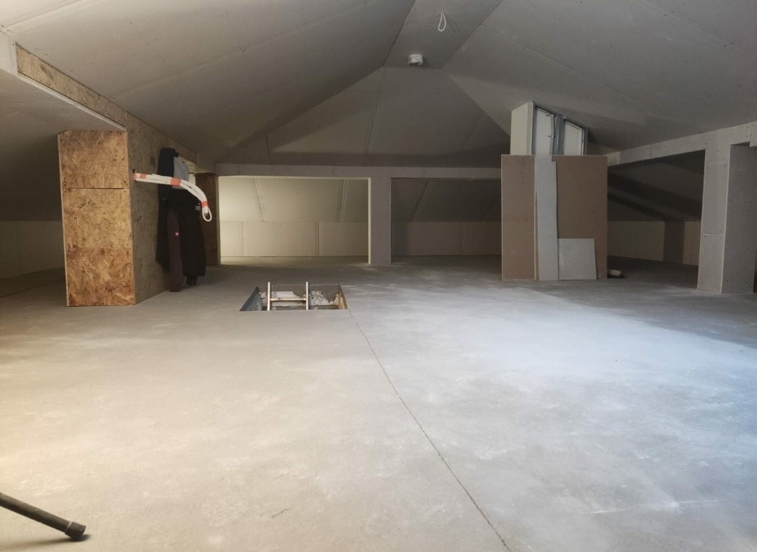 Poddasze w trakcie adaptacji: surowy betonowy podkład, ściany z płyt gipsowo-kartonowych, otwór w podłodze z metalową ramą, widoczne elementy konstrukcyjne dachu.