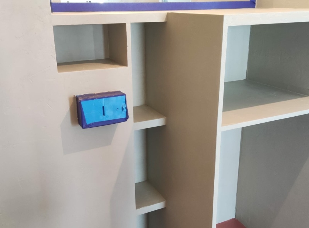 Niemiędzynarodowy regał z półkami wnękowymi, zabezpieczony taśmą malarską gniazdko elektryczne na ścianie, świeżo pomalowane na szaro wnętrze w trakcie prac wykończeniowych.