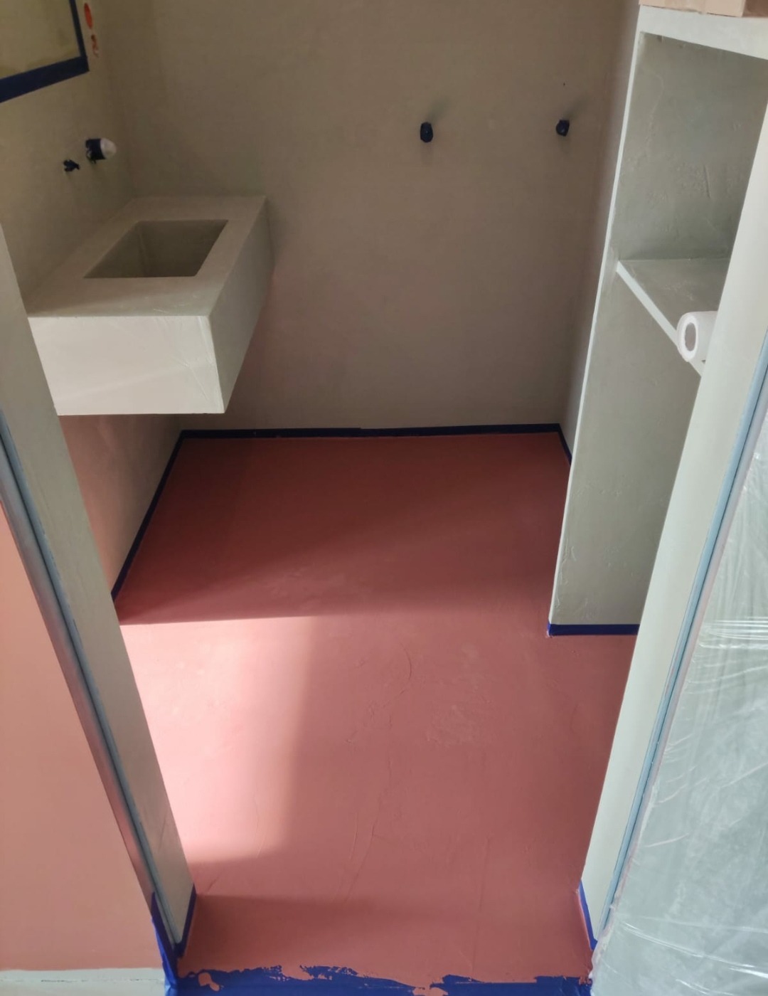 Łazienka w trakcie wykańczania, ściany i podłoga w odcieniach różu, zabezpieczone niebieską taśmą malarską, minimalistyczna umywalka i półki wnękowe.