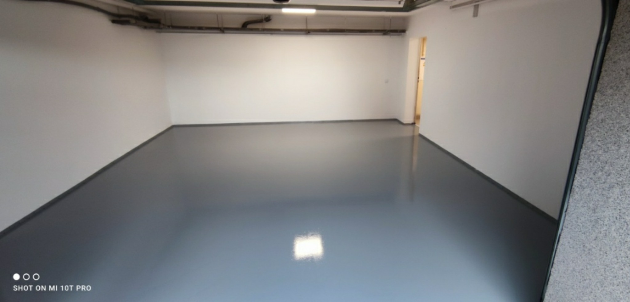 Szara, błyszcząca posadzka epoksydowa w pustym pomieszczeniu z białymi ścianami i widocznymi rurami pod sufitem.