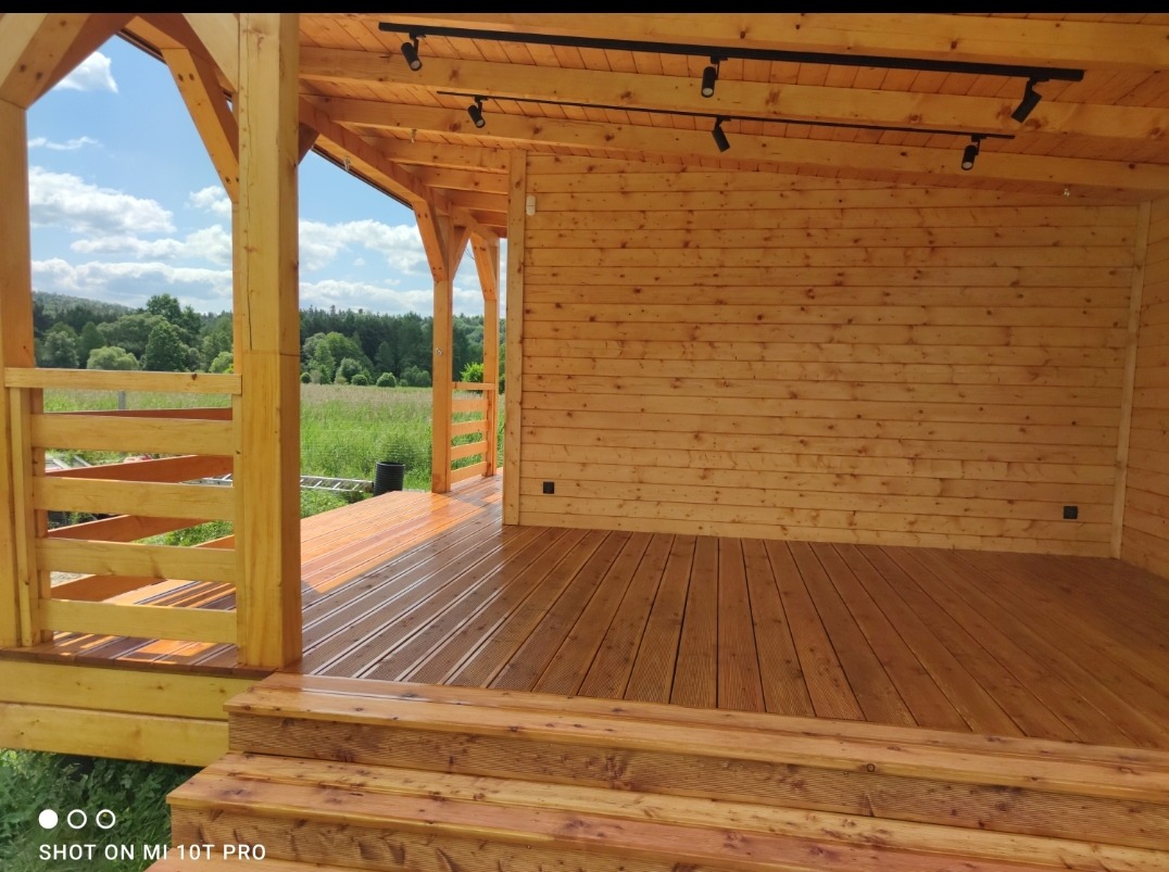 Drewniana altana z widokiem na zielone pole i las, wykończona w jasnym drewnie, z oświetleniem punktowym na suficie i schodami wejściowymi.