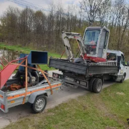 Transport mini-koparki Takeuchi TB016 na platformie samochodu dostawczego wraz z przyczepą ze zrębkowarką, widok z boku na tle zieleni.