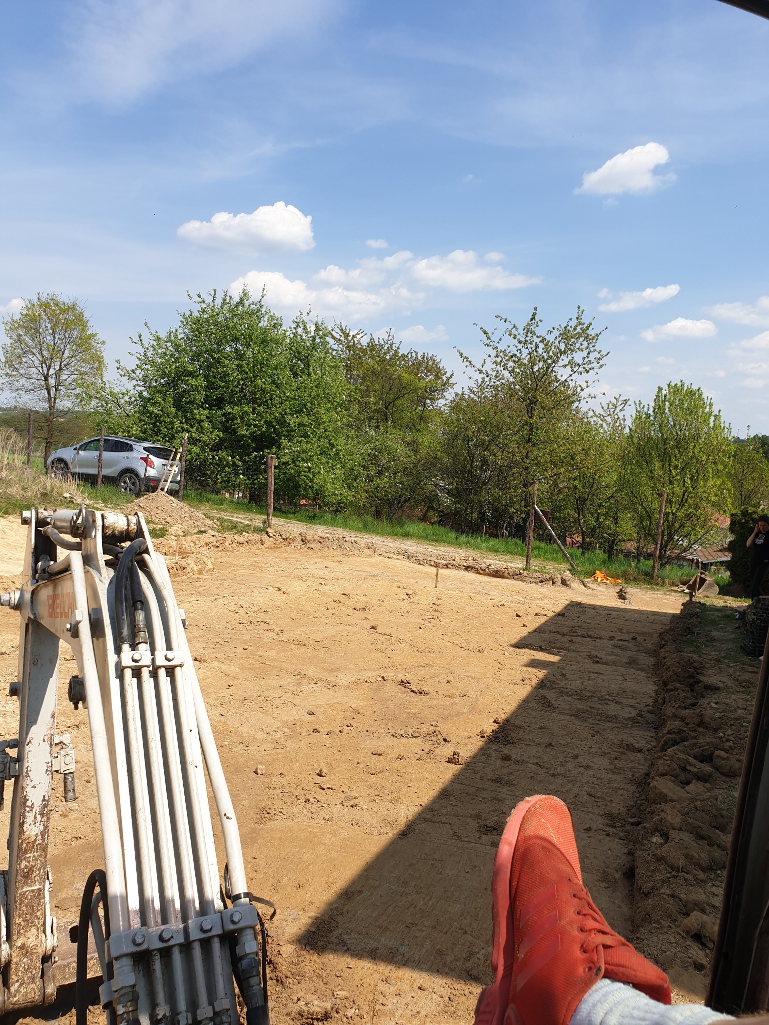 Widok z kabiny koparki na przygotowany teren pod wylanie fundamentów, widoczne oznaczenia geodezyjne, w tle drzewa i zaparkowany samochód.