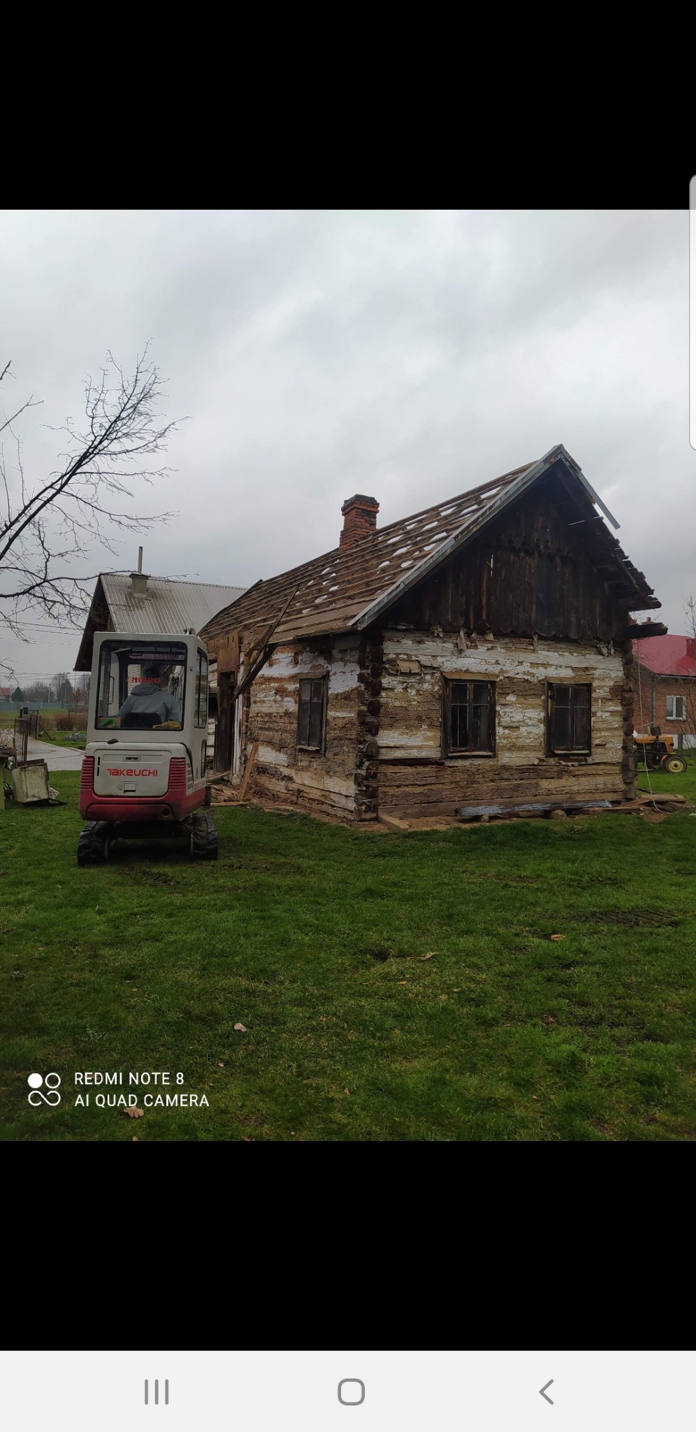 Mini koparka Takeuchi obok częściowo rozebranego, starego, drewnianego domu z odsłoniętą konstrukcją dachu na tle pochmurnego nieba.