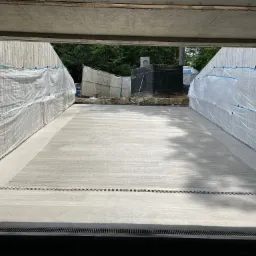 Świeżo wylana, gładka, betonowa posadzka z widocznymi szczelinami dylatacyjnymi i odpływem liniowym; zabezpieczona folią budowlaną na ścianach w trakcie prac.