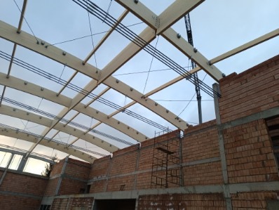 Wnętrze budowy hali z drewnianą konstrukcją dachu, widoczne ceglane ściany w trakcie wznoszenia oraz rusztowanie.