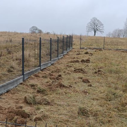 Długi, panelowy ogrodzenie z podmurówką betonową, rozciągające się przez pole z przygotowanym terenem pod dalsze prace, widoczne drzewa w tle pod pochmurnym niebem.
