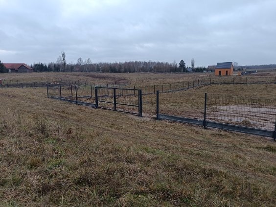 Nowo postawione, metalowe ogrodzenie panelowe na rozległym, jesiennym terenie z widocznymi budynkami w tle, sfotografowane w pochmurny dzień.