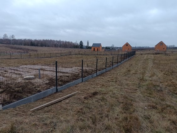 Długi, częściowo ukończony płot z metalowej siatki na betonowej podmurówce, biegnący przez pole w kierunku trzech drewnianych domów w tle, pochmurny dzień.