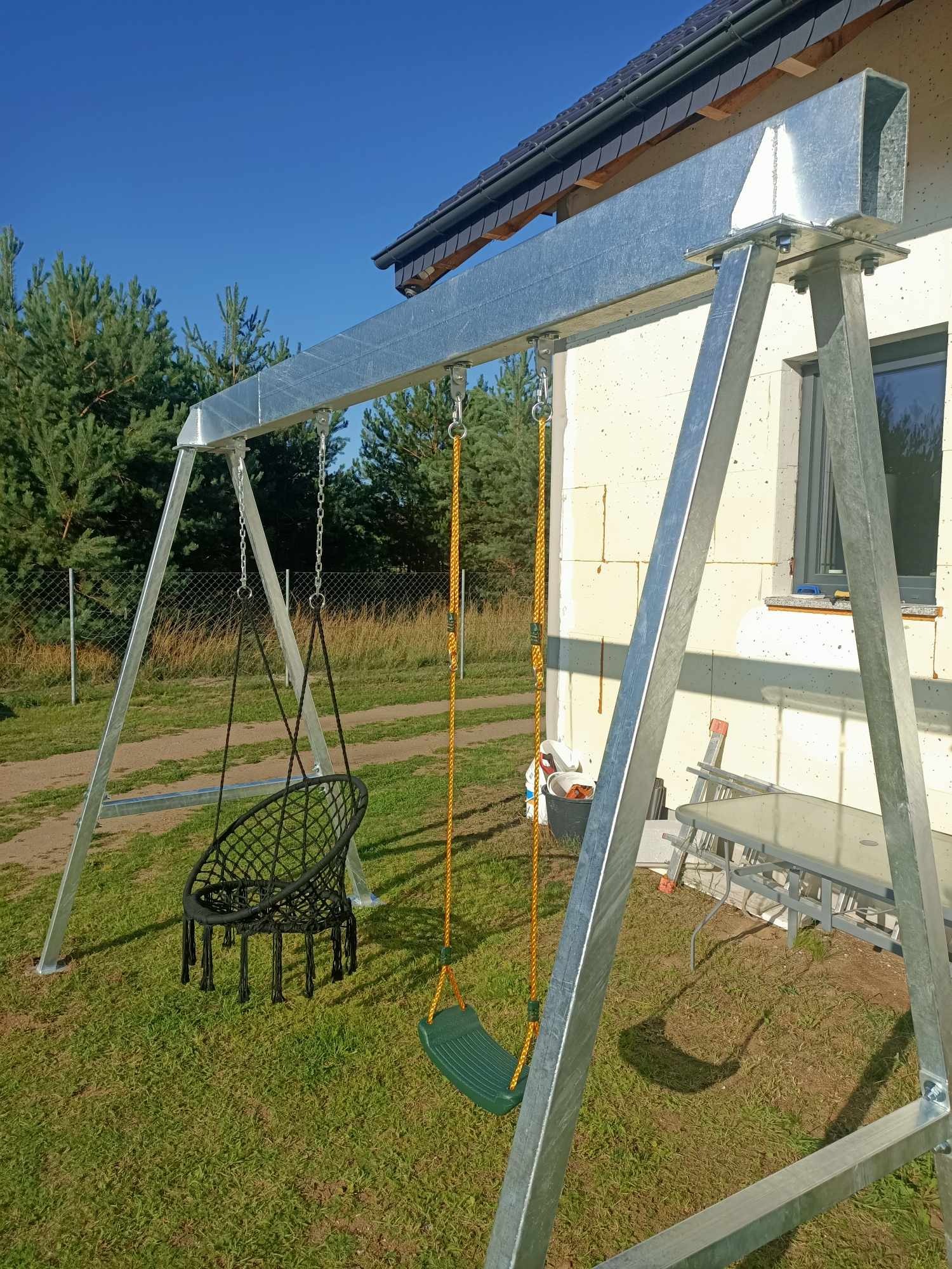 Metalowa konstrukcja huśtawki z dwoma siedziskami: czarnym, plecionym fotelem wiszącym i zielonym siedziskiem na żółtym sznurze, na tle domu w trakcie budowy i zielonego trawnika.