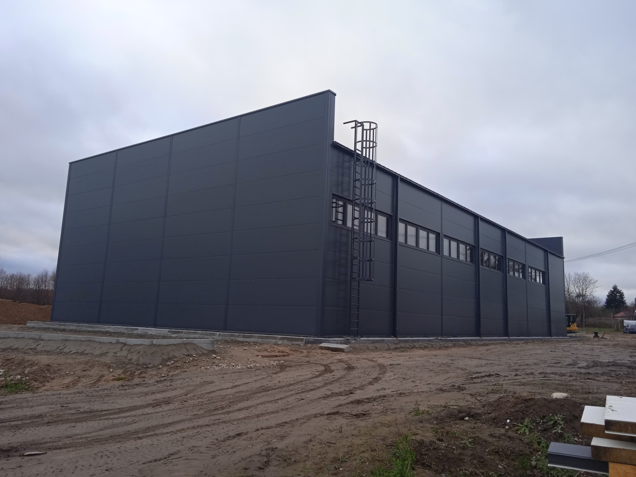 Nowoczesna, ciemnoszara hala przemysłowa z płyt warstwowych, z zewnętrzną drabinką po lewej stronie, widoczna na tle pochmurnego nieba i błotnistego terenu budowy.