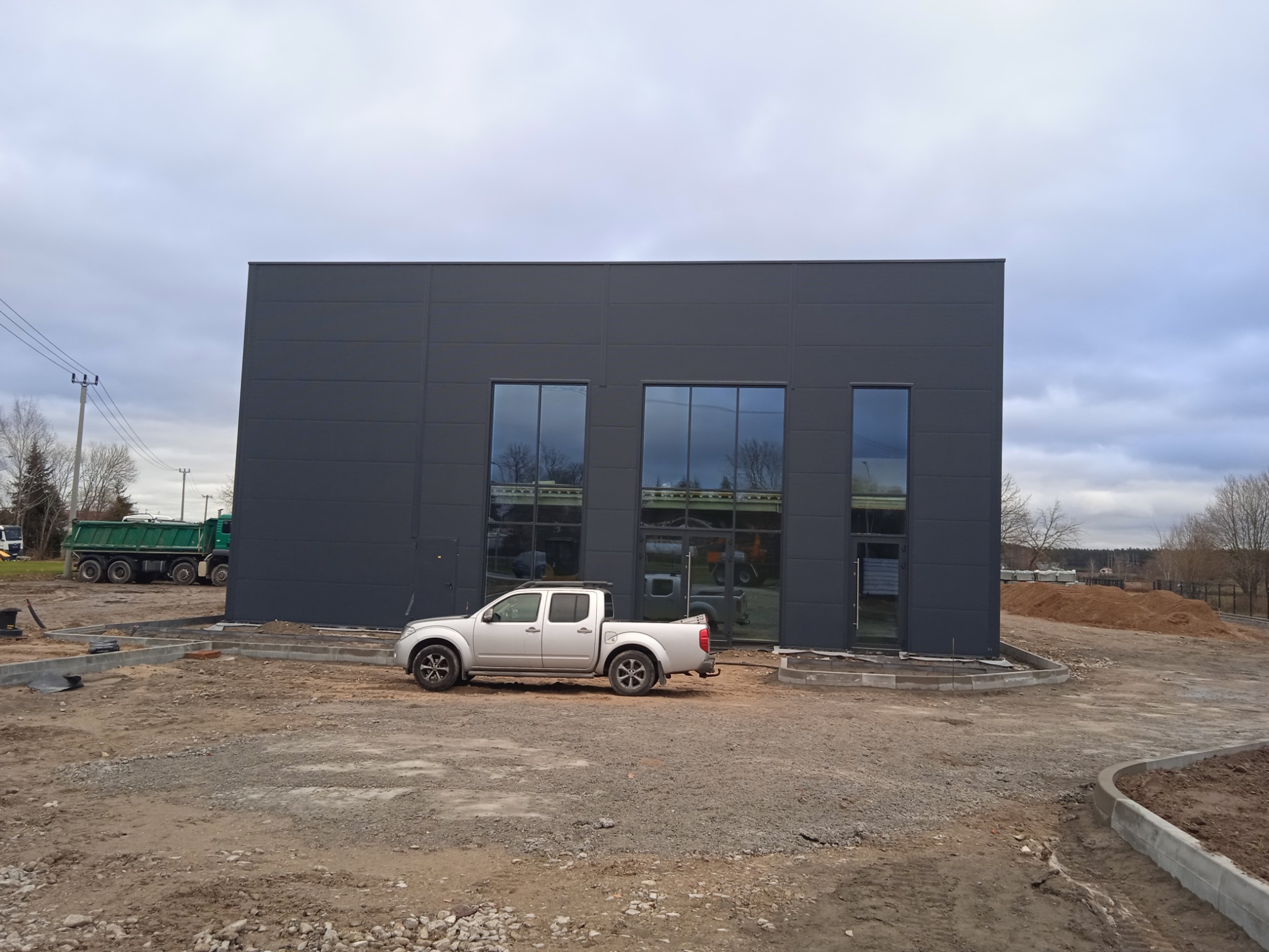 Nowo wybudowana hala z szarej płyty warstwowej z trzema oknami i srebrnym pickupem zaparkowanym przed nią, widoczny teren budowy i zielony wywrotka w tle.