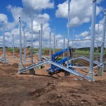 Montaż konstrukcji stalowej hali na betonowych fundamentach; widoczny podnośnik koszowy na placu budowy, błękitne niebo z chmurami w tle.