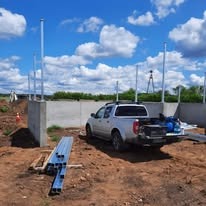 W trakcie budowy ogrodzenia: widoczne betonowe podmurówki, metalowe słupki, zaparkowany samochód typu pickup z narzędziami na pace, w tle zielone pola i błękitne niebo z chmurami.