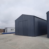 Szara hala magazynowa z blachy trapezowej, widok z zewnątrz, zamknięte duże drzwi przesuwne, widoczny fragment drugiej hali po prawej stronie i mniejszy budynek w tle, pochmurne niebo.