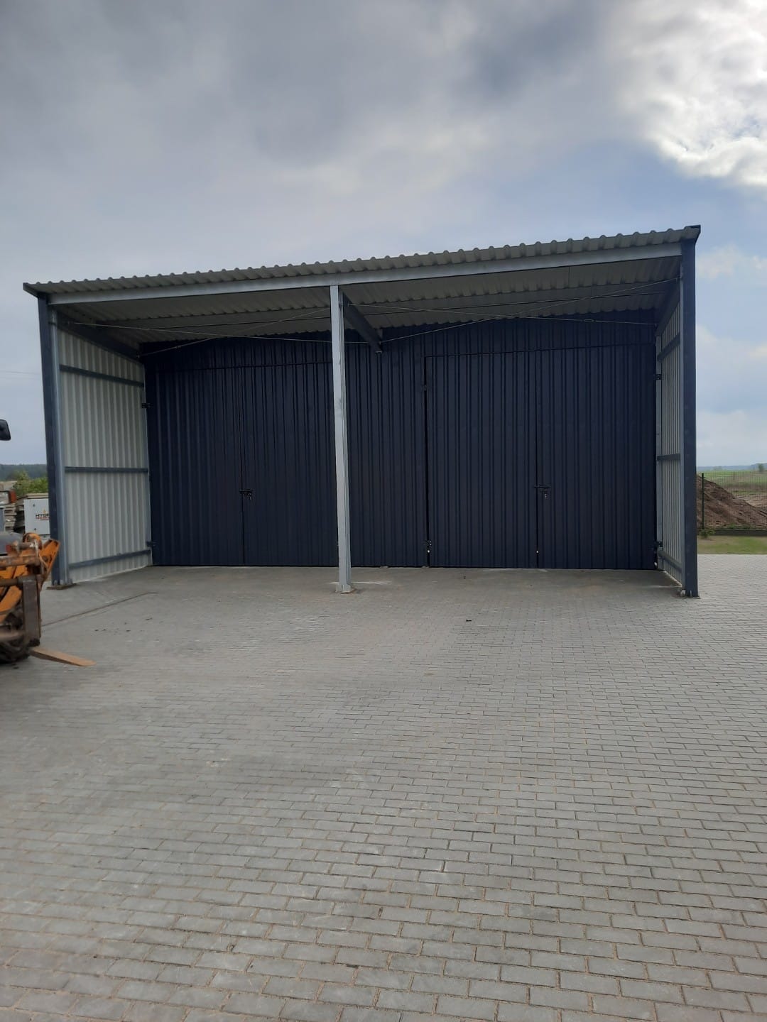 Gotowa hala z dwoma czarnymi, zamkniętymi bramami wjazdowymi, widoczna na tle pochmurnego nieba i brukowanej nawierzchni, z fragmentem pomarańczowego sprzętu budowlanego po lewej stronie.