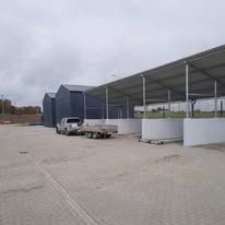 Nowoczesna hala magazynowa z szarej blachy trapezowej, widoczny fragment konstrukcji zadaszenia oraz zaparkowany pick-up z przyczepką na wybrukowanym placu manewrowym w pochmurny dzień.