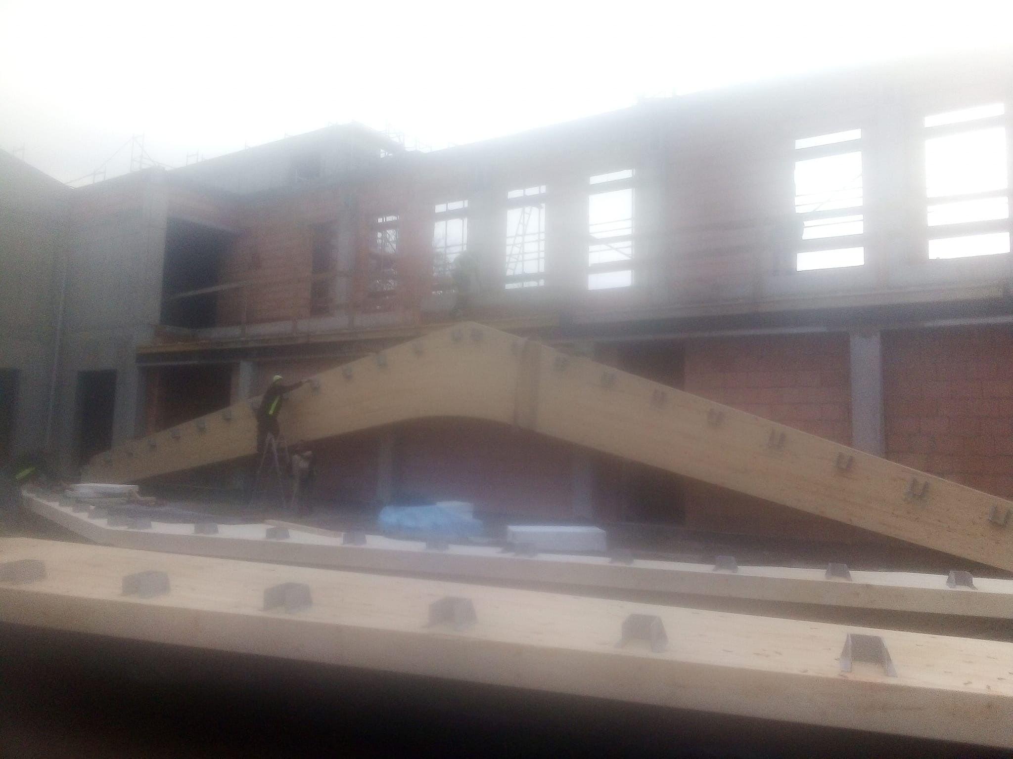 Konstrukcja hali w trakcie budowy: drewniane dźwigary z metalowymi łącznikami ustawione na placu budowy, w tle mur z cegły i pracujący robotnicy.