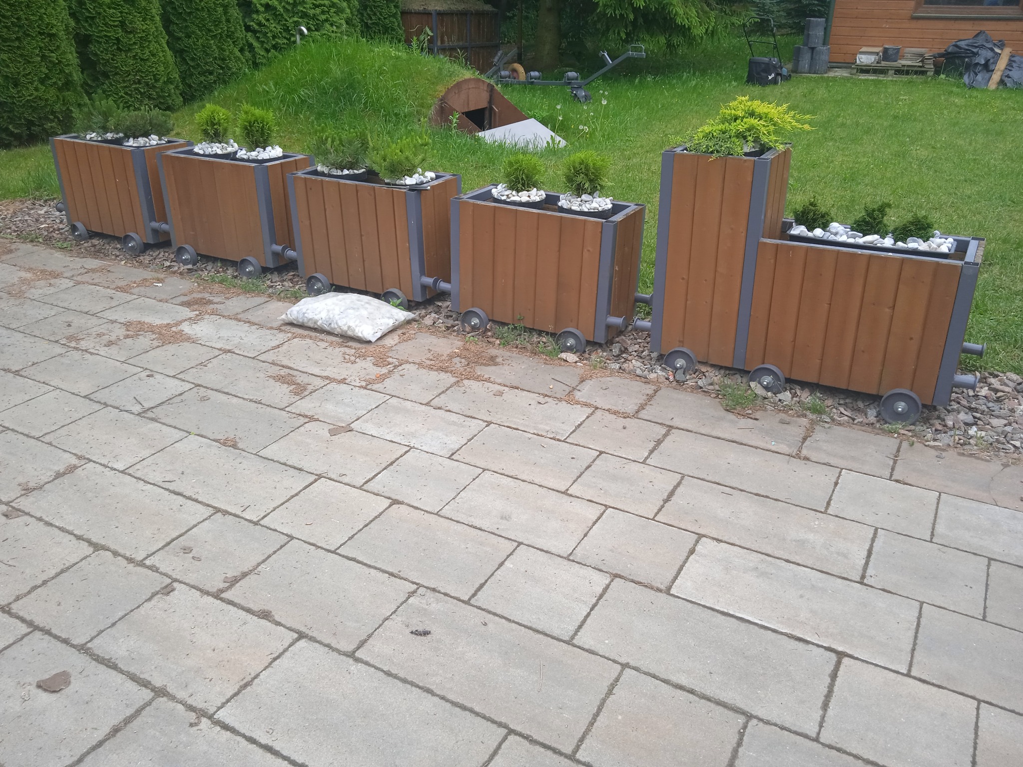 Dekoracyjne, drewniane donice w kształcie wagoników kolejowych z roślinami i białymi kamieniami, ustawione na kostce brukowej w ogrodzie.