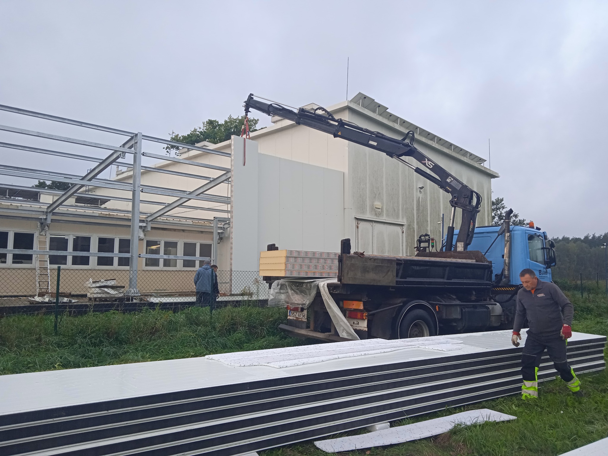 Montaż płyty warstwowej na stalowej konstrukcji hali przy użyciu dźwigu samochodowego; pracownik w odzieży roboczej kontroluje proces, a w tle widoczny budynek z oknami.