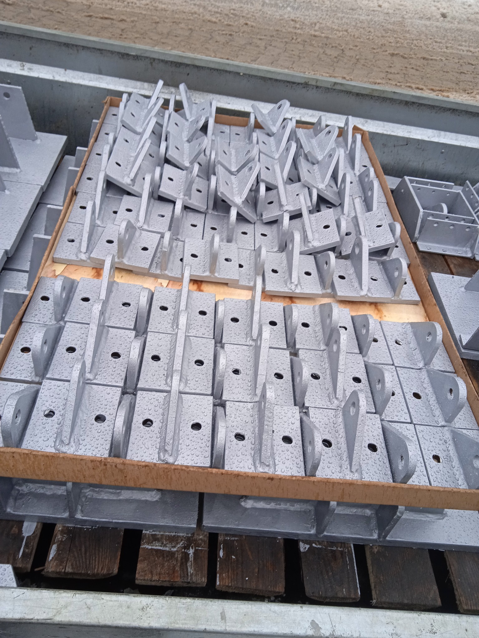 Szare, metalowe elementy konstrukcyjne w skrzyni transportowej, pokryte kroplami wody, ułożone równo w rzędach.