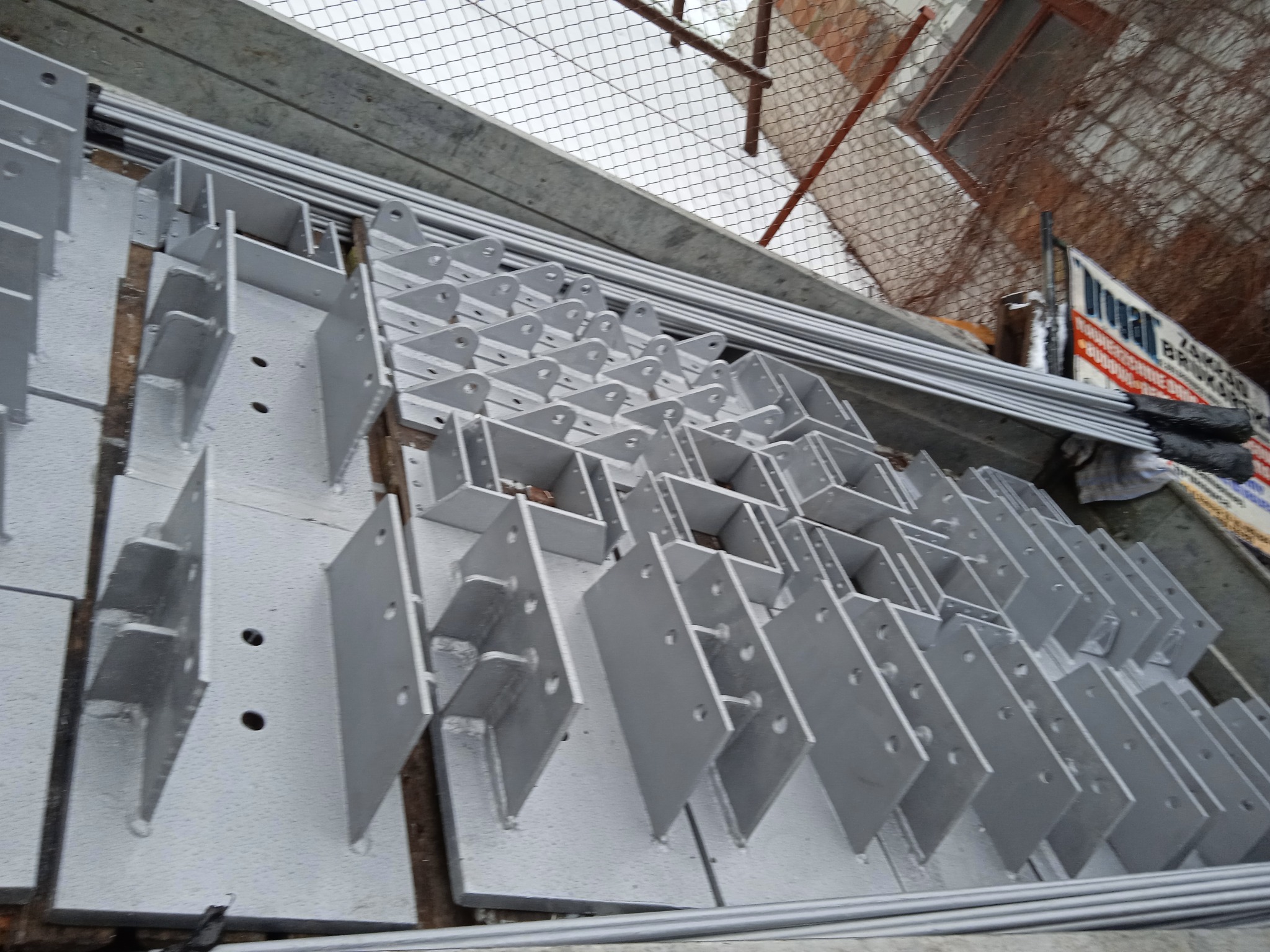 Stalowe, pomalowane na szaro elementy konstrukcyjne z otworami, ułożone w rzędach na zewnątrz, prawdopodobnie przygotowane do montażu.