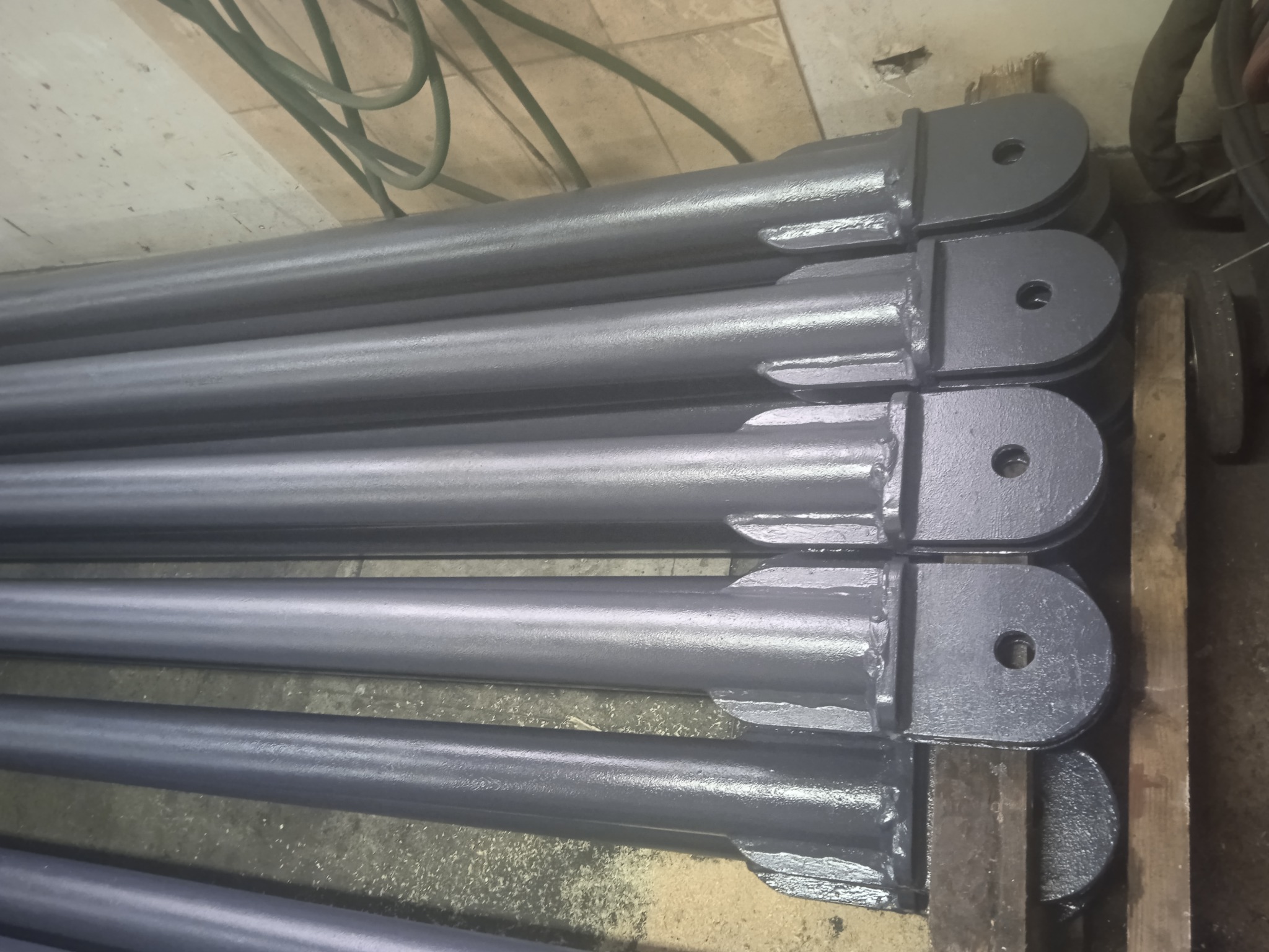 Stalowe, pomalowane na szaro słupy z przyspawanymi elementami montażowymi, ułożone równolegle na drewnianej powierzchni. Widoczne spawy i otwory montażowe.