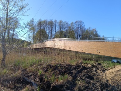 Drewniany most z metalową barierką, łagodny łuk, otoczony zielenią. Ujęcie z dołu, w tle drzewa i błękitne niebo, ziemia z odkopaną darnią na pierwszym planie.