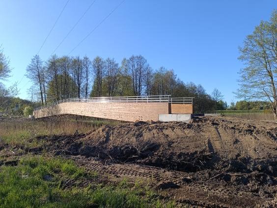 Drewniany mostek z metalową barierką, wkomponowany w krajobraz z zielenią i ziemią. Element konstrukcji stalowej, widoczne słupy wysokiego napięcia w tle.