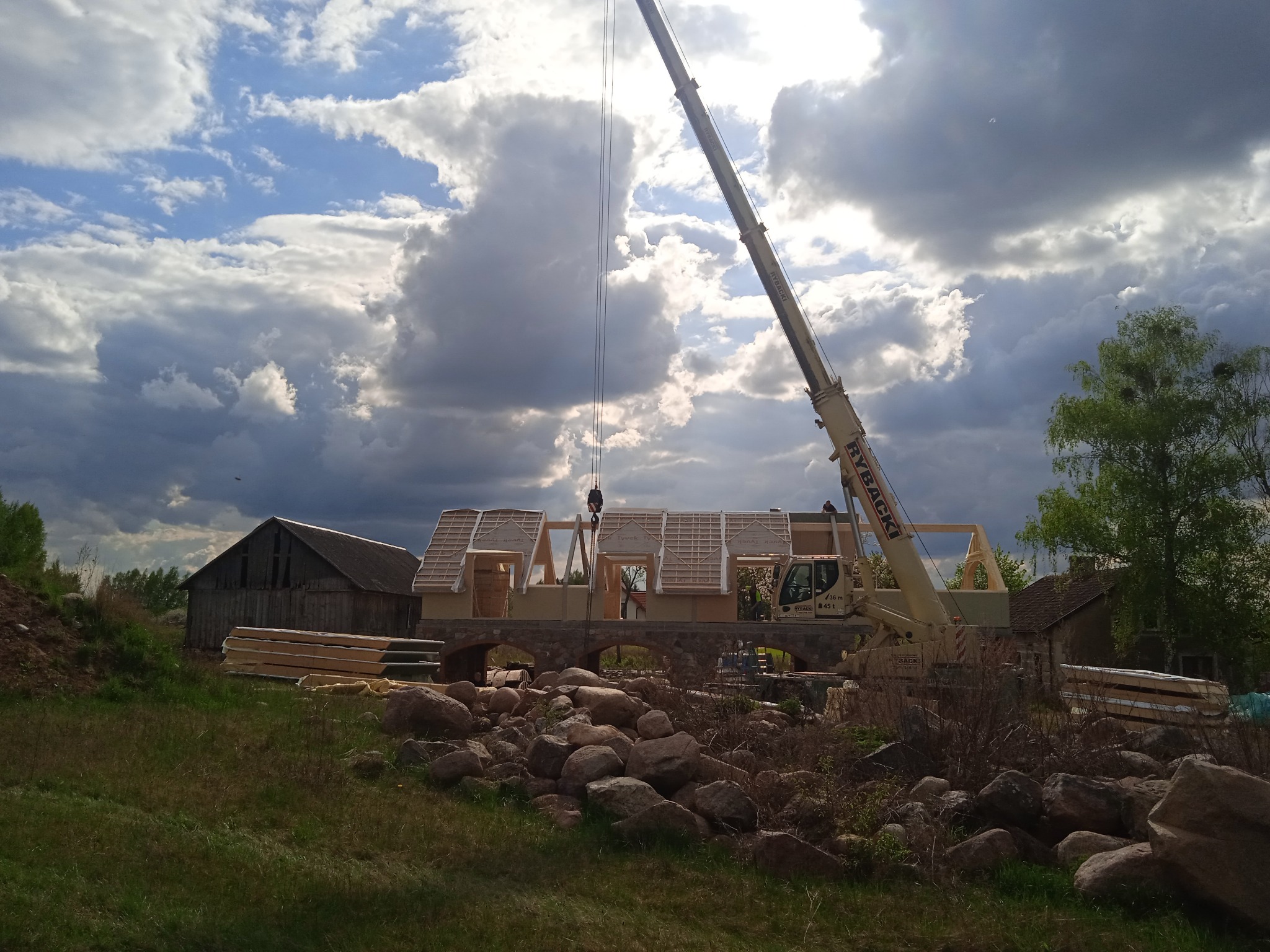 Budowa domu szkieletowego z użyciem dźwigu na tle pochmurnego nieba. W tle widoczny stary budynek gospodarczy i ułożone płyty warstwowe.