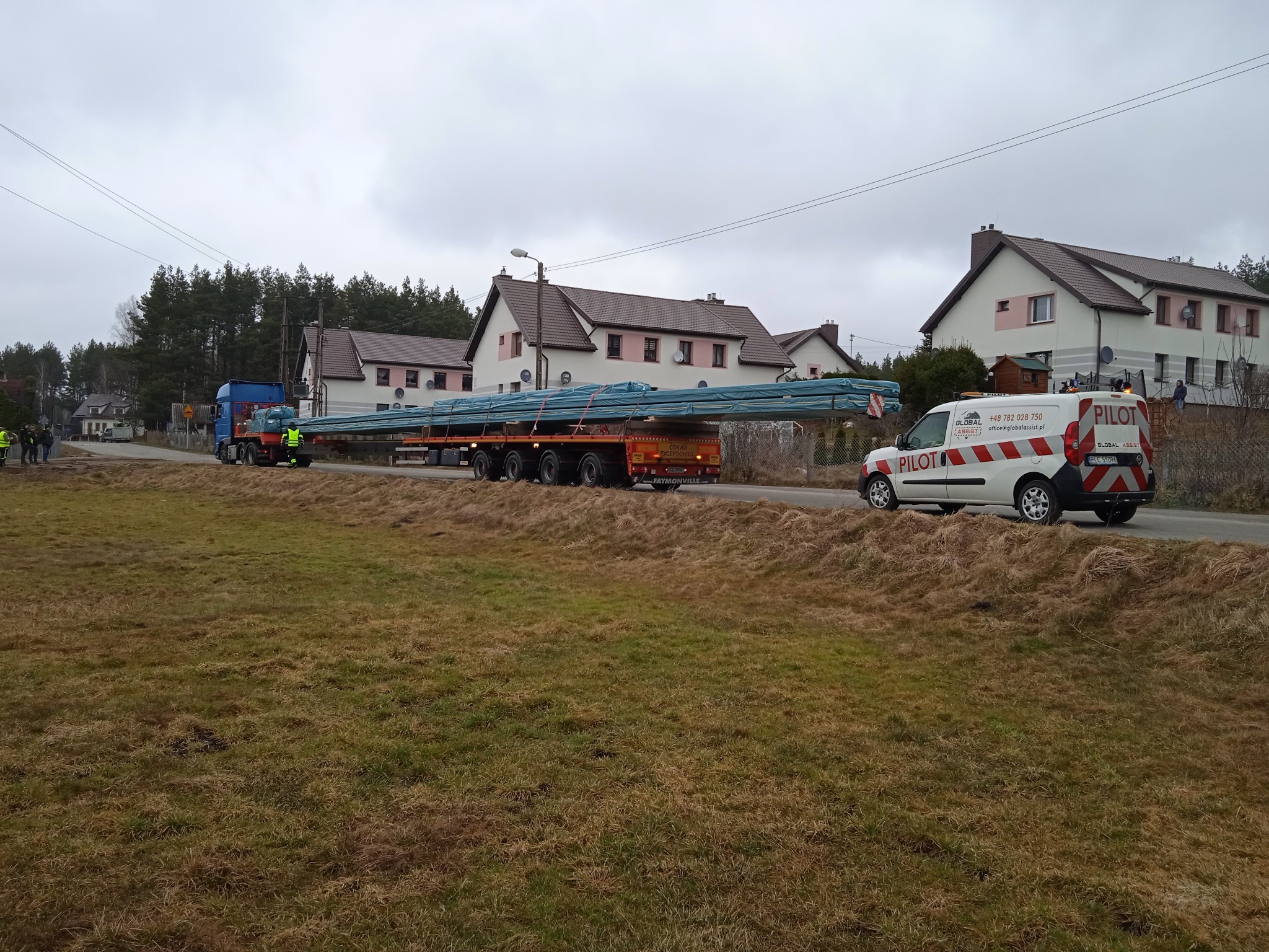 Transport długich, niebieskich elementów konstrukcyjnych na naczepie ciężarówki, eskortowany przez pojazd pilotujący na drodze w wiejskiej scenerii.