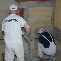 Dwóch mężczyzn w roboczych ubraniach aplikuje tynk maszynowy na ścianę z siatką wzmacniającą; jeden stoi z wężem, drugi klęczy i wygładza tynk