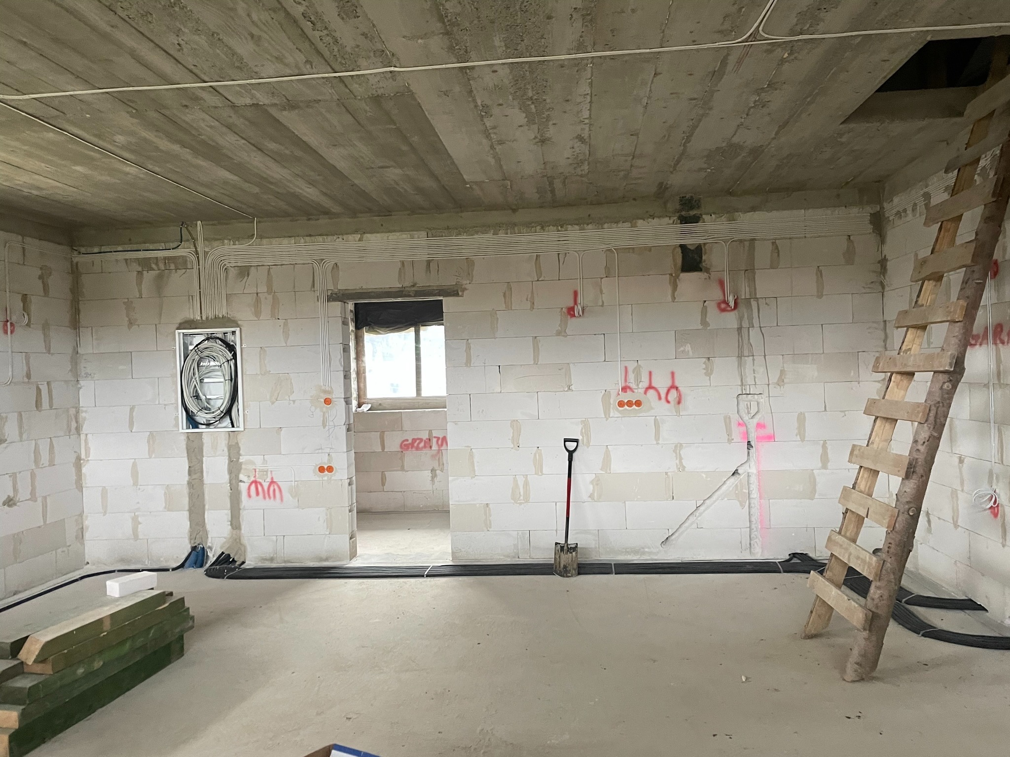 Wnętrze surowego budynku w trakcie prac elektrycznych: widoczne kable prowadzone po ścianach, otwory pod gniazdka z naniesionymi oznaczeniami, skrzynka z przewodami, drewniana drabina oparta...