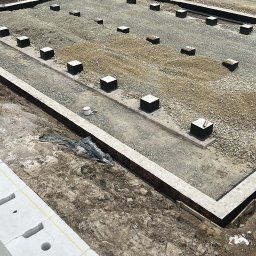 Budownictwo Mateusz Kubik - Rozległy plac budowy z przygotowanymi fundamentami: prostokątne betonowe ławy i liczne kwadratowe stopy na podsypce żwirowej. Widoczne wykopy z izolacją i materiały budowlane.