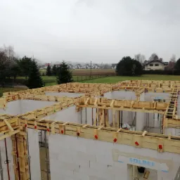 Wznoszenie ścian domu z betonu komórkowego z drewnianym szalunkiem, widok z góry na budowę w pochmurny dzień, w tle zabudowa jednorodzinna.