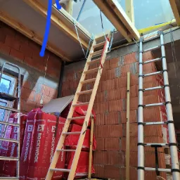 Montaż schodów strychowych w surowym stanie, widoczne ocieplenie wełną mineralną Rockwool, ceglana ściana, drabina aluminiowa i poziomica.