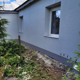 A-Z PIOTR-BUD - Świeżo pomalowana, szara elewacja budynku z dwoma oknami, widoczny fragment dachu, w tle błękitne niebo z chmurami, wokół budynku widoczne resztki gruzu i roślinność.