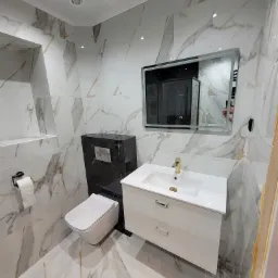 Nowoczesna łazienka wykończona płytkami imitującymi marmur, z podwieszaną toaletą w zabudowie z czarnego marmuru, białą szafką z umywalką i złotą baterią oraz podświetlanym lustrem.