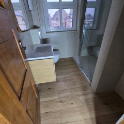 A-Z PIOTR-BUD - Jasna łazienka z drewnianą podłogą i szafką pod umywalką. Widok na ulicę przez okno. Kabina prysznicowa ze szklaną ścianą. Drewniane drzwi częściowo otwarte.