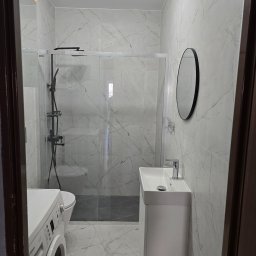 A-Z PIOTR-BUD - Nowoczesna łazienka z kabiną prysznicową typu walk-in, umywalką na szafce i pralką. Płytki imitujące marmur na ścianach i podłodze. Okrągłe lustro.