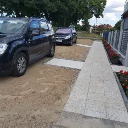 Ułożona ścieżka z jasnych płyt chodnikowych na podsypce piaskowej, z dwoma zaparkowanymi samochodami i rabatami kwiatowymi wzdłuż ogrodzenia.