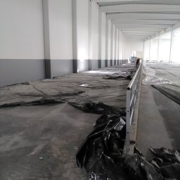 A-Z PIOTR-BUD - Wnętrze hali przemysłowej w trakcie przygotowań do prac wykończeniowych, podłoga zabezpieczona czarną folią, widoczne metalowe słupy konstrukcyjne i fragment rusztowania w oddali.