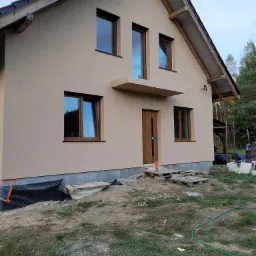 Nowo otynkowany dom jednorodzinny z beżową elewacją, brązowymi oknami i drzwiami, widoczne palety i teren w trakcie zagospodarowywania.