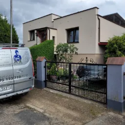 Nowoczesny dom jednorodzinny z jasną elewacją, widoczny zza czarnej, ozdobnej bramy wjazdowej. Na podjeździe stoi srebrny bus firmy budowlanej z logo i numerem telefonu.