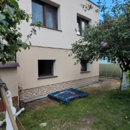 Dwukondygnacyjny dom z jasną elewacją, widoczne okna z brązowymi ramami, fragment ogrodu z drzewami i plandeką, dolna część elewacji wykończona imitacją kamienia.